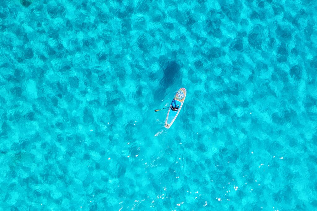 Stand-up-Paddler auf kristallklarem türkisfarbenem Wasser der Balearen, von oben fotografiert – mediterranes Urlaubsparadies für Segelreisen.