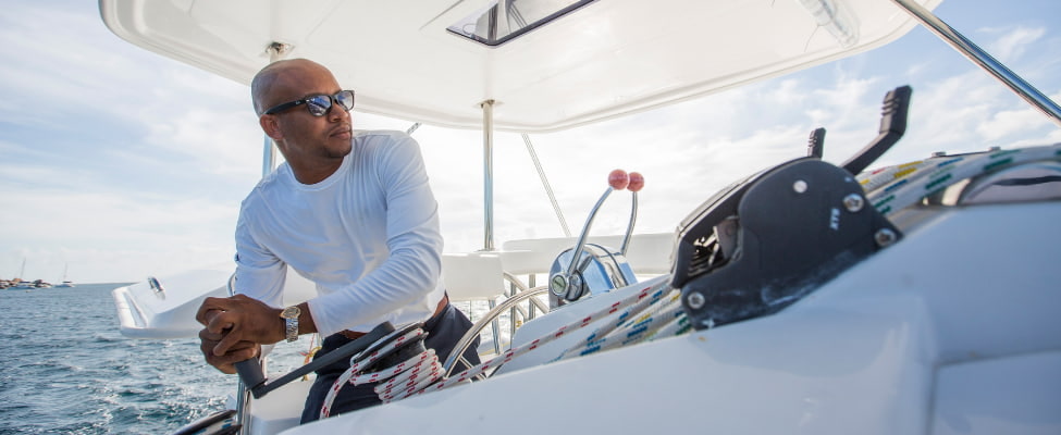 Ein erfahrener Skipper mit Sonnenbrille steuert eine Segelyacht über das offene Meer. Im Hintergrund glitzert das tiefblaue Wasser unter der Sonne, während der Blick des Skippers konzentriert nach vorne gerichtet ist – ein Symbol für Sicherheit, Erfahrung und entspannte Chartertörns mit professioneller Begleitung.