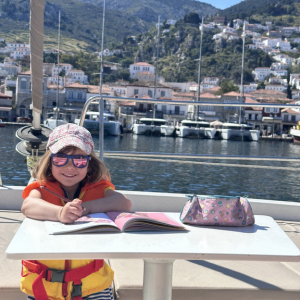 Ein Kind sitzt an einem kleinen Tisch an Bord eines Bootes und malt oder schreibt konzentriert in ein aufgeschlagenes Heft. Es trägt eine orangefarbene Schwimmweste sowie eine helle Kappe mit Muster und eine Sonnenbrille mit getönten Gläsern, die das Gesicht vor der Sonne schützt. Das Kind lächelt leicht und wirkt entspannt und zufrieden bei seiner Beschäftigung. Auf dem Tisch liegt neben dem Heft ein Federmäppchen mit buntem Design. Im Hintergrund erstreckt sich ein malerischer Hafen mit mehreren festgemachten Booten und Yachten, dahinter steigen dicht bebaute Häuser an einem grünen Hügel hinauf. Die Szene vermittelt eine Mischung aus Urlaubsstimmung, Kreativität und entspanntem Leben am Wasser.