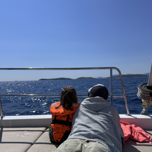 Zwei Personen liegen bäuchlings nebeneinander auf dem Deck eines Bootes und schauen über die Reling hinaus auf das tiefblaue Meer. Eine Person trägt eine orangefarbene Schwimmweste, die andere einen grauen Pullover und eine Kappe. Die Wasseroberfläche glitzert in der Sonne, und am Horizont zeichnen sich sanfte Hügel oder Inseln ab. Die Szene vermittelt Ruhe und Neugier, als würden beide gemeinsam die Weite des Meeres genießen.
