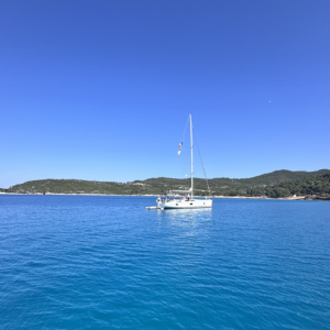 Ein einzelnes Segelboot liegt ruhig auf einem türkisblauen, klaren Meer. Das Wasser ist so still, dass sich das Boot leicht darin spiegelt. Im Hintergrund erstreckt sich eine grüne, hügelige Küstenlandschaft unter einem wolkenlosen, strahlend blauen Himmel. Die Atmosphäre wirkt friedlich und beinahe zeitlos, als wäre das Boot fernab von jeglicher Hektik unterwegs.