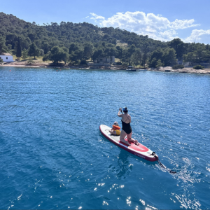 Eine Person steht auf einem Stand-up-Paddle-Board und paddelt über das klare, tiefblaue Wasser. Vor ihr sitzt ein Kind, beide tragen Schwimmwesten. Die Umgebung zeigt eine ruhige Bucht mit bewaldeten Hügeln und wenigen Gebäuden entlang der Küste. Das Wasser glitzert im Sonnenlicht, und die Szene vermittelt ein Gefühl von Freizeit, Abenteuer und gemeinsamer Aktivität in der Natur.