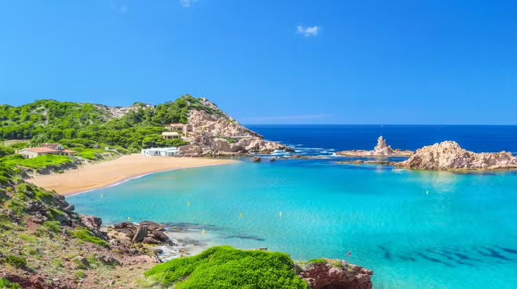 türkisfarbene Badebucht der Balearen in Menorca vor einer felsigen Küstenlandschaft mit kristallklarem Mittelmeer. Die geschützte Ankerbucht bietet ideale Bedingungen für Yachtcharter auf Menorca und einen exklusiven Segelurlaub entlang der malerischen Inselküsten der Balearen unter strahlend blauem Himmel.