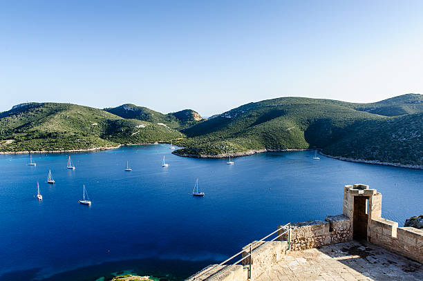 Blick auf die Insel Cabrera mit tiefblauem Mittelmeer, zahlreichen Segelbooten und grünen Felslandschaften im Hintergrund. Rechts unten ist ein Teil einer historischen Steinmauer-Festung zu sehen – ideales Revier für Segelreisen und Yachtcharter auf den Balearen mit GlobeSailor.